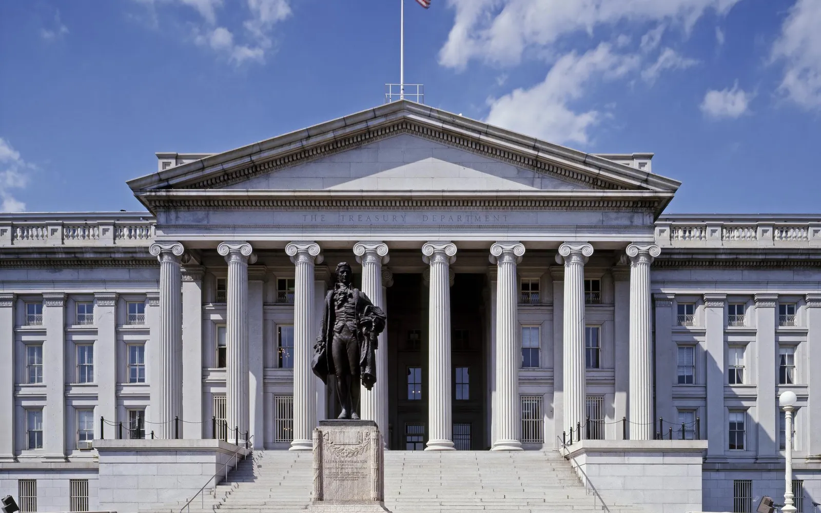 The columned south front of the U.S. Treasury Department building in Washington under a blue sky, with a bronze statue in front