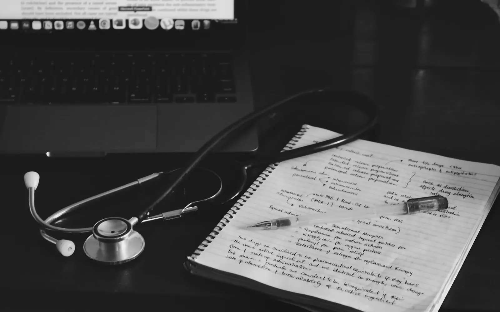 A stethoscope resting on handwritten notes beside a laptop, in black and white