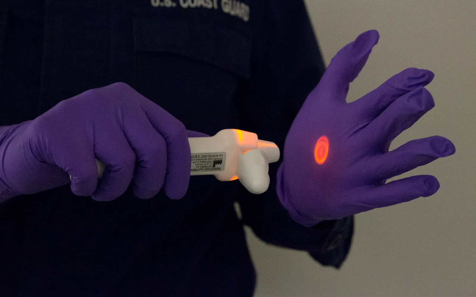 A U.S. Coast Guard medic in purple gloves holds a handheld infrared thermometer during enhanced Ebola screening at JFK airport in 2014