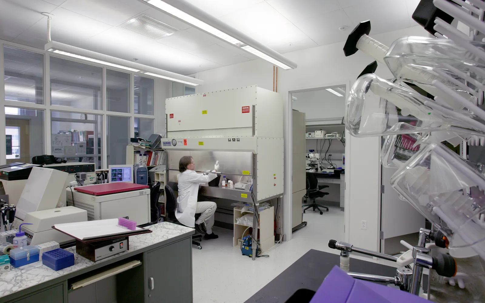 A scientist in a white coat works at a biosafety cabinet in an FDA life sciences lab, with glassware in the foreground
