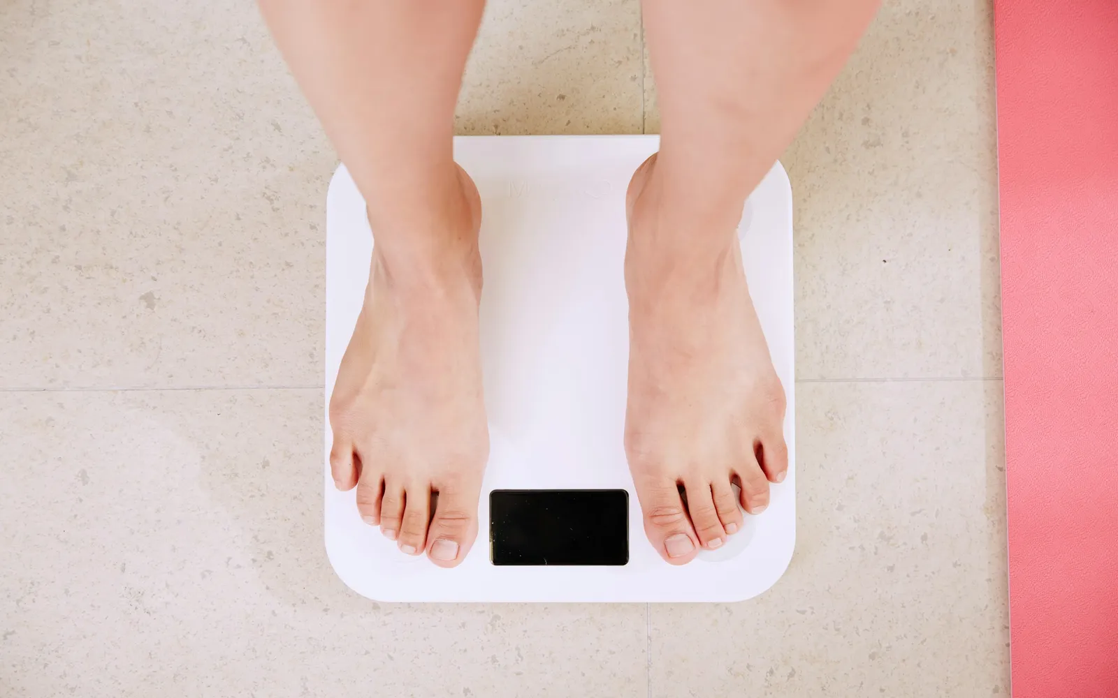 Bare feet on a white digital bathroom scale on a stone floor, beside a pink exercise mat