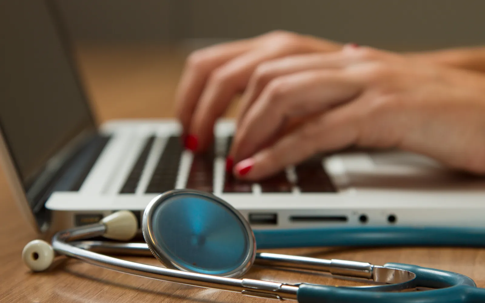 Hands typing on a laptop with a stethoscope lying in the foreground
