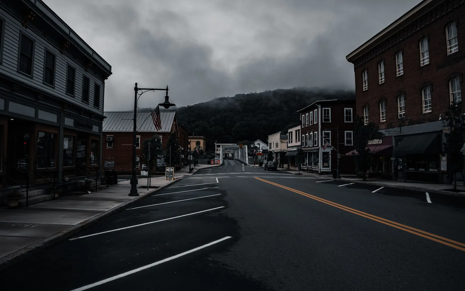 An empty main street in a small town with brick storefronts, under low clouds and a wooded hill