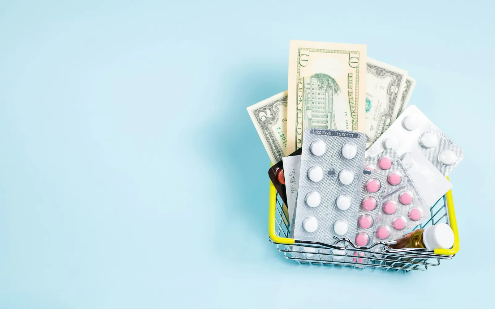 A small wire shopping basket holding blister packs of pills, a pill bottle and dollar bills, on a pale blue background