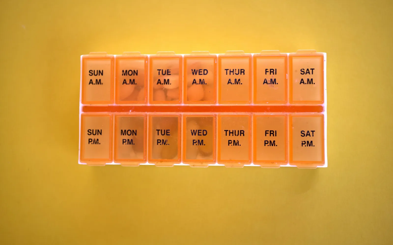 An orange weekly pill organizer with morning and evening compartments for each day, on a yellow background
