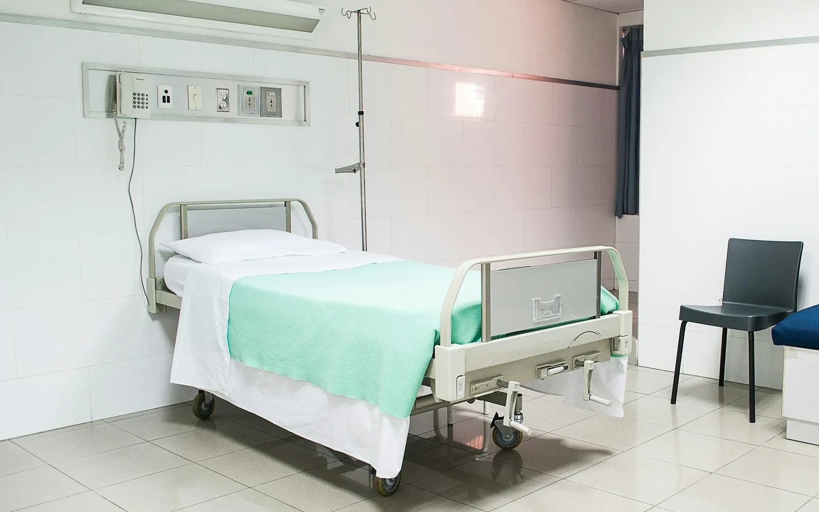 An empty hospital bed with side rails and a green blanket in a white tiled room