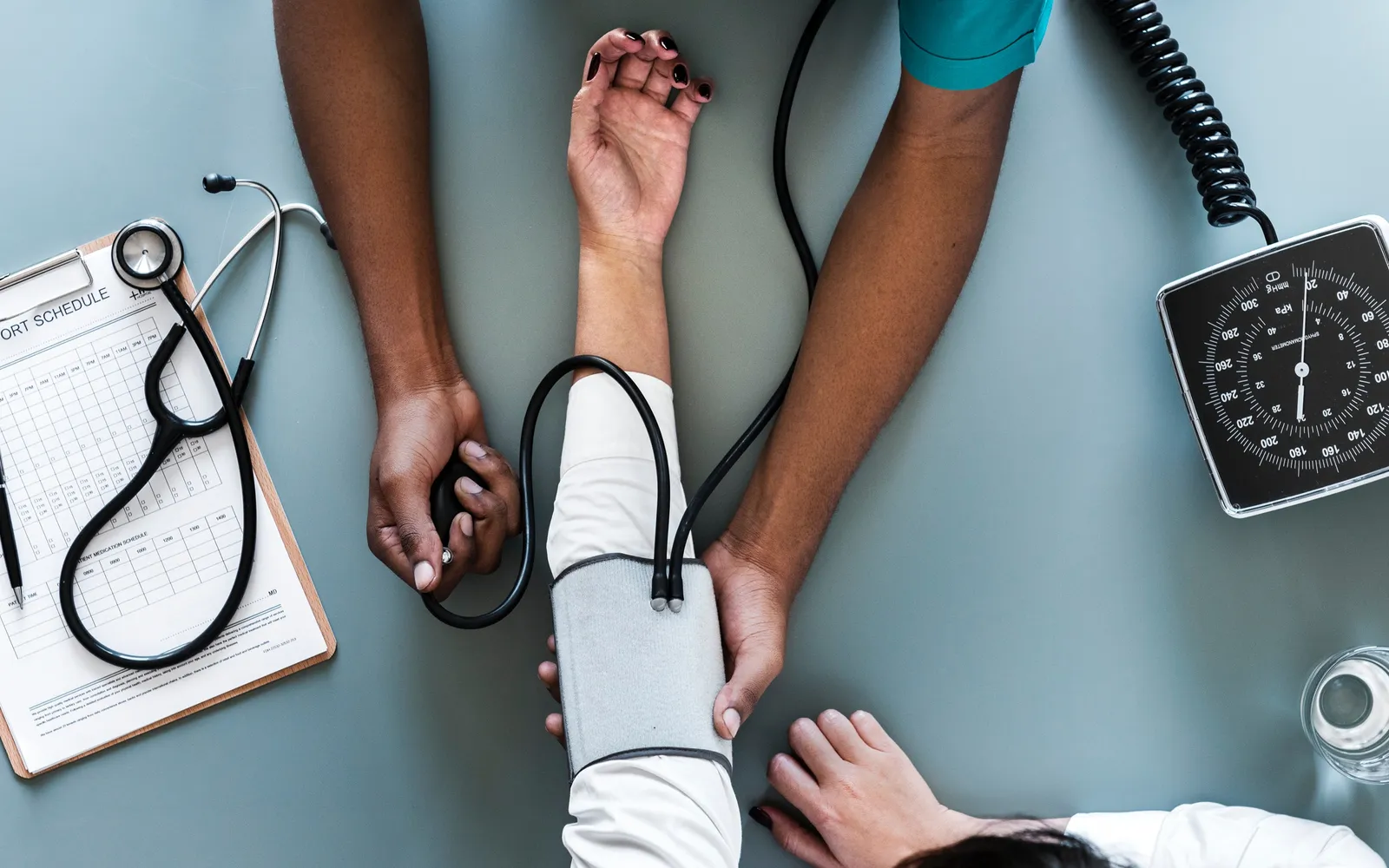 Seen from above on a blue gray table, a clinician's hands wrap a blood pressure cuff on a patient's arm beside a dial gauge, stethoscope and clipboard